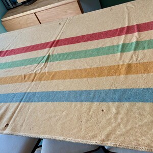 May include: A rectangular cream-colored wool blanket featuring horizontal stripes in red, green, yellow, and blue. The blanket has a woven texture, frayed edges, and a small rectangular fabric patch sewn onto the bottom right corner with visible text.