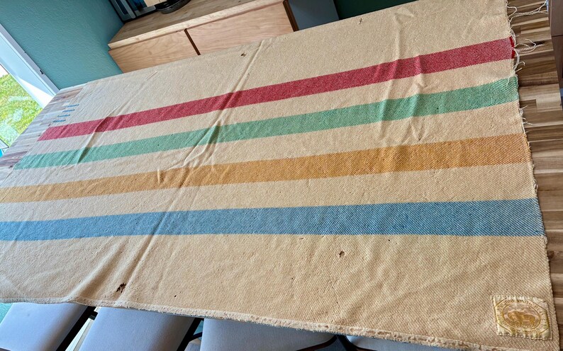 May include: A rectangular cream-colored wool blanket featuring horizontal stripes in red, green, yellow, and blue. The blanket has a woven texture, frayed edges, and a small rectangular fabric patch sewn onto the bottom right corner with visible text.