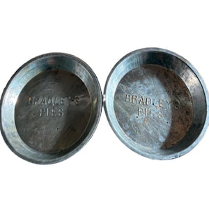 May include: Two round, silver-colored metal pie tins with a weathered finish. Each tin features the embossed text "BRADLEY'S PIES" clearly visible in the center of the base. The edges are raised and smooth, showing signs of use and oxidation.
