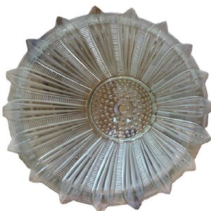 May include: A circular clear glass ceiling light fixture featuring a radial sunburst pattern of elongated, pointed petals extending from a central circular disc textured with small, raised dots and a single hole in the center.