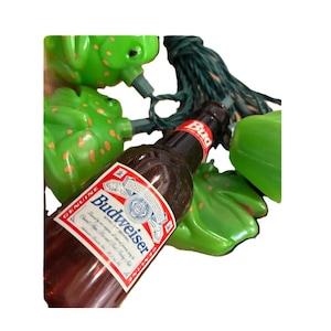 May include: A brown Budweiser beer bottle with a red and white label, and green frog-shaped Christmas lights. The lights are connected by a green wire. The bottle is partially visible.