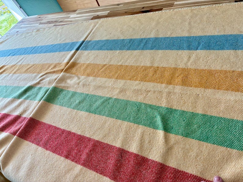 May include: A woven blanket featuring a cream-colored base with horizontal stripes in shades of light blue, yellow, green, and red. The fabric has a visible textured weave pattern throughout, creating a consistent and uniform appearance across the surface.