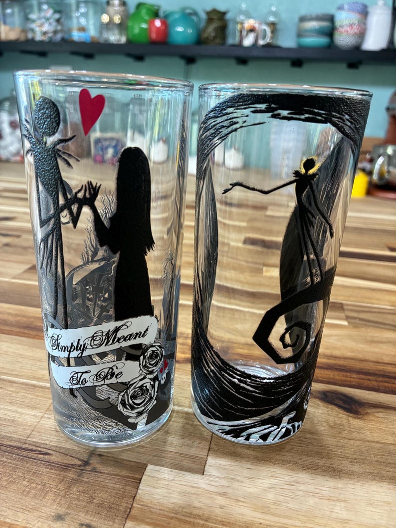 Puede incluir: Dos vasos de cristal transparente con ilustraciones de siluetas negras. Uno muestra dos figuras tomadas de la mano con un coraz&oacute;n rojo y el texto Simply Meant To Be. El otro vaso muestra una figura solitaria dentro de un marco negro giratorio.