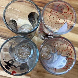Puede incluir: Cuatro vasos de vidrio transparente vistos desde arriba. Dos vasos presentan siluetas de personajes en negro, mientras que los otros dos muestran patrones de enredaderas rojas y un rostro de personaje blanco. Tienen forma cil&iacute;ndrica.