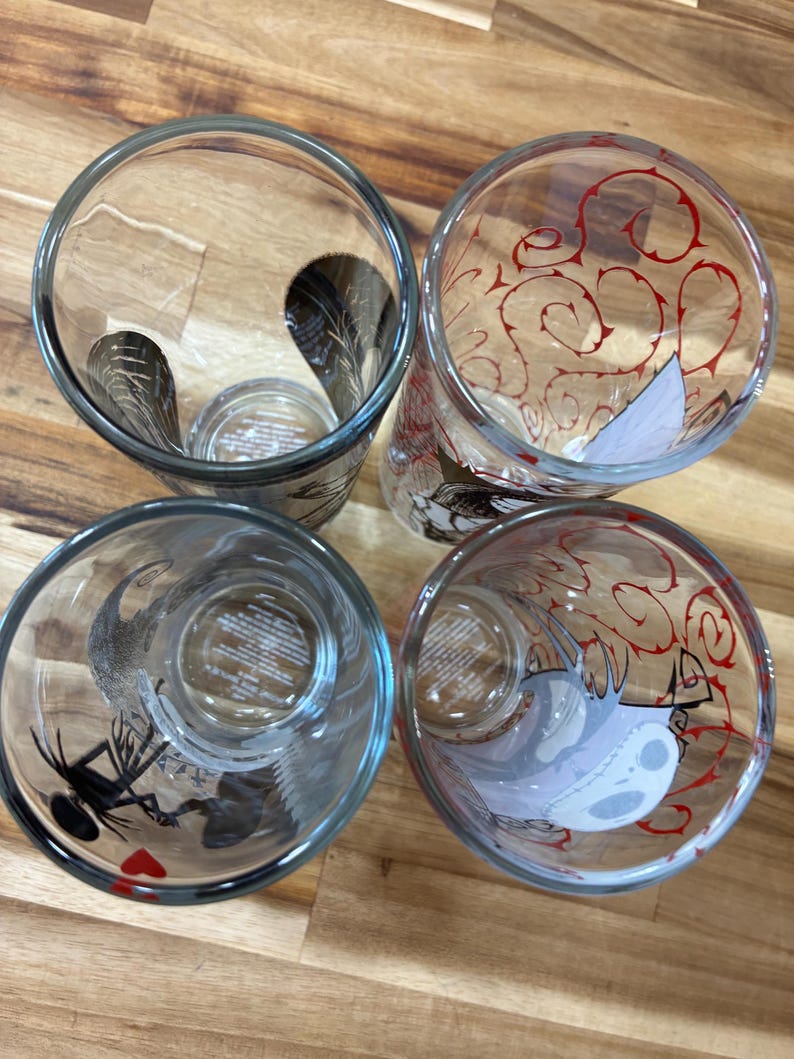 Puede incluir: Cuatro vasos de vidrio transparente vistos desde arriba. Dos vasos presentan siluetas de personajes en negro, mientras que los otros dos muestran patrones de enredaderas rojas y un rostro de personaje blanco. Tienen forma cil&iacute;ndrica.