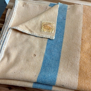 May include: A woven blanket featuring a light yellow base with a prominent vertical blue stripe and a wider orange stripe. The blanket has a folded corner revealing a square fabric label with a circular emblem and text, and shows small surface wear.