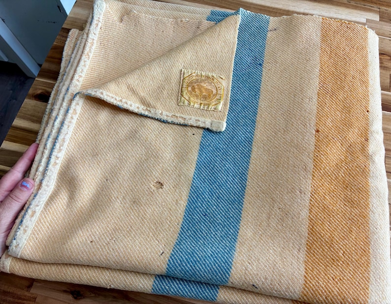 May include: A woven blanket featuring a light yellow base with a prominent vertical blue stripe and a wider orange stripe. The blanket has a folded corner revealing a square fabric label with a circular emblem and text, and shows small surface wear.