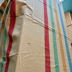 May include: A woven textile blanket featuring a cream-colored base with vertical stripes in red, green, yellow, and blue. The fabric displays a visible texture and several small holes scattered across the surface, indicating wear and age.