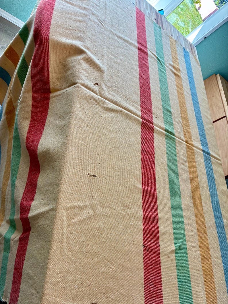 May include: A woven textile blanket featuring a cream-colored base with vertical stripes in red, green, yellow, and blue. The fabric displays a visible texture and several small holes scattered across the surface, indicating wear and age.