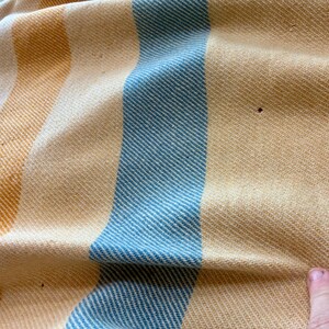 May include: A woven textile featuring vertical stripes in shades of light blue, soft yellow, and cream. The fabric has a distinct diagonal twill weave pattern throughout. There are small, dark, circular imperfections visible on the yellow sections.