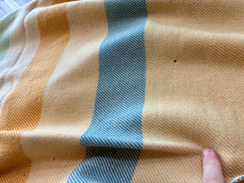 May include: A woven textile featuring vertical stripes in shades of light blue, soft yellow, and cream. The fabric has a distinct diagonal twill weave pattern throughout. There are small, dark, circular imperfections visible on the yellow sections.
