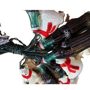 May include: String of Christmas lights featuring Coca-Cola bottles and polar bear figures. The bottles are dark brown with white "Coca-Cola" text. The polar bears are white with red scarves and the Coca-Cola logo.