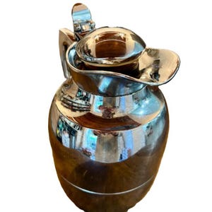 May include: A silver-plated pitcher with a wooden base and lid. The pitcher has a curved handle and a spout. The surface of the pitcher reflects the surrounding environment. The pitcher is likely used for serving beverages.