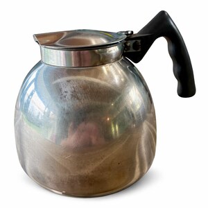 May include: A stainless steel coffee pot with a black handle and lid. The pot has a rounded body and a shiny, reflective surface. The handle is attached to the side of the pot, and the lid sits on top.