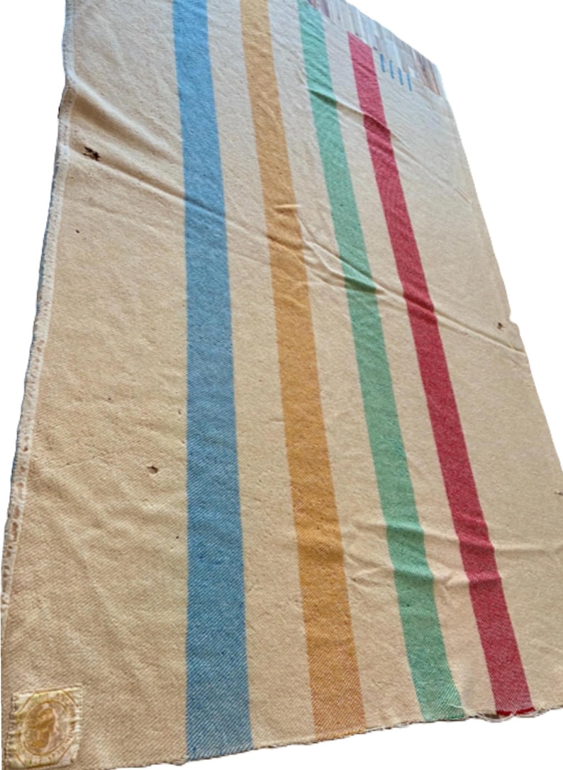 May include: A rectangular off-white woven blanket featuring four vertical stripes in light blue, yellow, green, and red. The fabric has a textured appearance with a small square label attached to the bottom left corner and visible frayed edges.