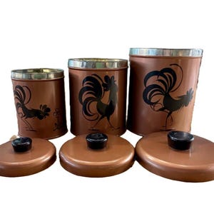 May include: A set of three copper-colored canisters with black rooster silhouettes. Each canister has a silver rim and a matching copper lid with a black knob. The canisters are of varying sizes, suitable for kitchen storage.