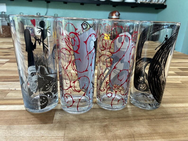 Puede incluir: Cuatro vasos de vidrio transparente con ilustraciones de personajes en blanco y negro y patrones de enredaderas rojas. Los dise&ntilde;os incluyen siluetas de una figura alta y delgada y una figura femenina, una cara esquel&eacute;tica y l&iacute;neas negras.