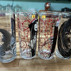 Puede incluir: Cuatro vasos transparentes con ilustraciones de personajes en blanco y negro y patrones de enredaderas rojas. Los dise&ntilde;os incluyen una figura esquel&eacute;tica alta, una mu&ntilde;eca de trapo cosida y una forma de luna creciente con l&iacute;neas.