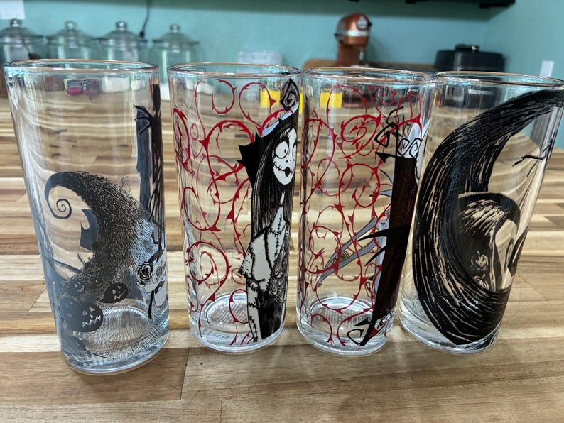 Puede incluir: Cuatro vasos transparentes con ilustraciones de personajes en blanco y negro y patrones de enredaderas rojas. Los dise&ntilde;os incluyen una figura esquel&eacute;tica alta, una mu&ntilde;eca de trapo cosida y una forma de luna creciente con l&iacute;neas.