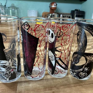 Puede incluir: Cuatro vasos de vidrio transparente con ilustraciones gr&aacute;ficas en negro y rojo de personajes de Pesadilla antes de Navidad. Un vaso muestra el texto "Meant to Be" con detalles de rosas, mientras que los otros presentan figuras esquel&eacute;ticas y enredaderas.