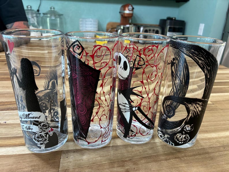 Puede incluir: Cuatro vasos de vidrio transparente con ilustraciones gr&aacute;ficas en negro y rojo de personajes de Pesadilla antes de Navidad. Un vaso muestra el texto "Meant to Be" con detalles de rosas, mientras que los otros presentan figuras esquel&eacute;ticas y enredaderas.