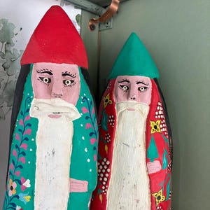 May include: Two hand-painted wooden gnome figures. One gnome wears a red hat and a green robe with floral patterns. The other gnome has a green hat and a red robe with yellow floral accents. Both have white beards and painted faces.