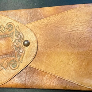 May include: A vintage leather wallet in shades of tan and brown. The front flap features an embossed decorative design with a central rectangular shape and ornate floral details. A small, dark metal button secures the flap.