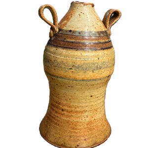 May include: A tall, hand-thrown ceramic vase with a speckled beige and tan finish. The vessel features two arched handles on the upper sides and horizontal bands in shades of brown and muted blue encircling the neck and body of the piece.