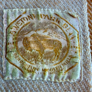 May include: A rectangular fabric label with a cream background and gold embroidery featuring a central bison illustration. The text reads "SUNSTONE PARK BLANKET" and "PENDLETON WOOLEN MILLS" with "PORTLAND OREGON" visible at the bottom of the circular emblem.