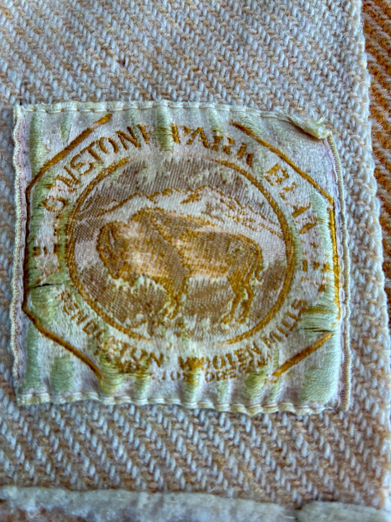 May include: A rectangular fabric label with a cream background and gold embroidery featuring a central bison illustration. The text reads "SUNSTONE PARK BLANKET" and "PENDLETON WOOLEN MILLS" with "PORTLAND OREGON" visible at the bottom of the circular emblem.