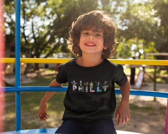 May include: A young child wearing a black t-shirt with a colourful graphic design that spells out "PHILLY". The child is sitting on a playground structure.