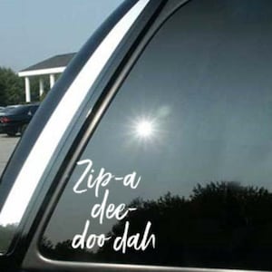 May include: A car window with a white decal that reads "Zip-a dee-doo dah" in a cursive font. The window is tinted, and the sun is reflecting off the glass. The car is parked outside.