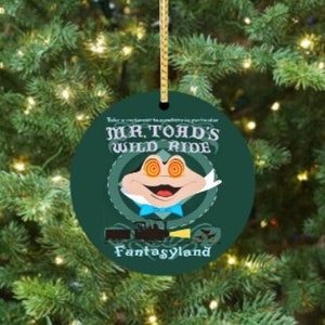 May include: A round, dark green Christmas ornament with the text "Mr. Toad's Wild Ride" and a cartoon toad illustration. The ornament also features a train graphic and the word "Fantasyland". It hangs from a gold string.
