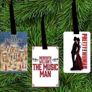 May include: Three rectangular Christmas ornaments with black ribbon hangers. The ornaments feature artwork from the musicals "My Fair Lady", "The Music Man", and "Pretty Woman". The "My Fair Lady" ornament shows a street scene, while the others display show logos.