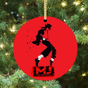 May include: Red round Christmas ornament featuring a black silhouette of a dancer in a hat and the text "MJ THE MUSICAL". The ornament hangs from a gold string against a backdrop of a Christmas tree.