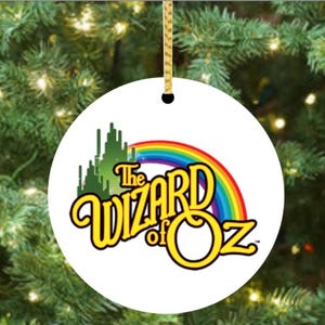 May include: Round white Christmas ornament featuring the text "The Wizard of Oz" in yellow and a rainbow. The ornament also includes a green illustration of the Emerald City. A gold string is attached for hanging.