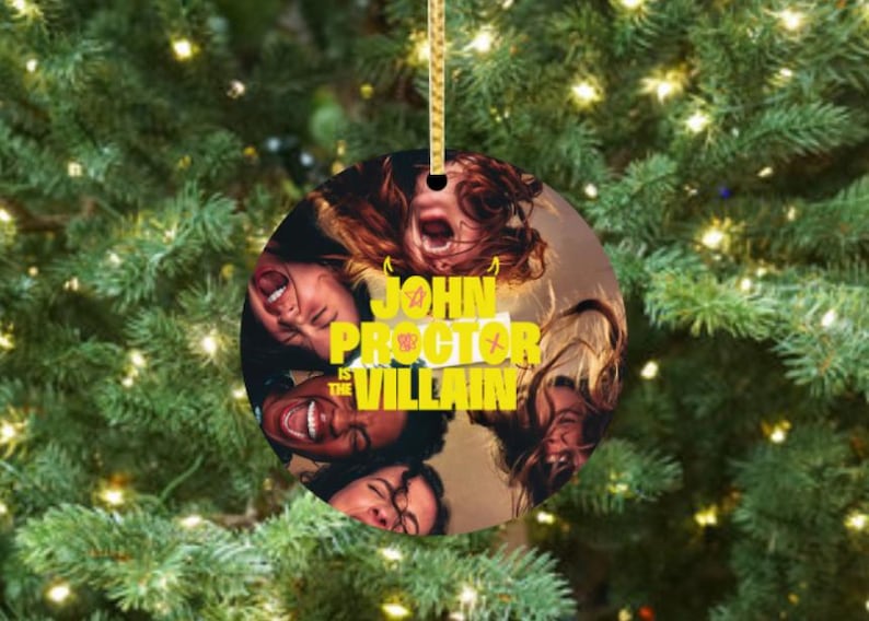 Puede incluir: Adorno navide&ntilde;o redondo con una cuerda dorada, que presenta un collage de caras con la boca abierta. El texto "JOHN PROCTOR is the VILLAIN" est&aacute; en amarillo. El adorno cuelga frente a un &aacute;rbol de Navidad.