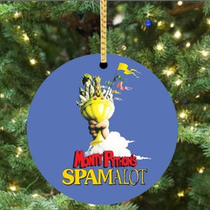 May include: Round Christmas ornament with a blue background featuring a yellow chalice held by a hand, topped with figures and flags. The text "Monty Python's SPAMALOT" is displayed in red and yellow.