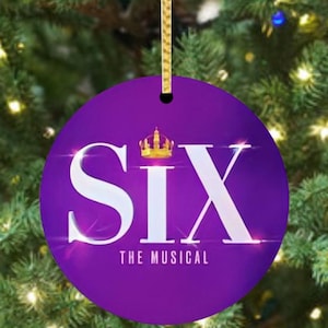 May include: A round, purple Christmas ornament with the word "SIX" in large white letters and a gold crown above. The text "THE MUSICAL" is below. The ornament hangs from a gold cord against a blurred Christmas tree background.