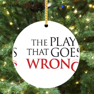 May include: A white, round Christmas ornament with the text "THE PLAY THAT GOES WRONG" in black and red. The ornament hangs from a gold string against a backdrop of a Christmas tree.