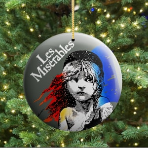 Puede incluir: Adorno navideño redondo con un diseño del musical "Les Misérables". El adorno tiene un fondo gris con el título en texto blanco. La imagen muestra a una joven con detalles rojos y azules.