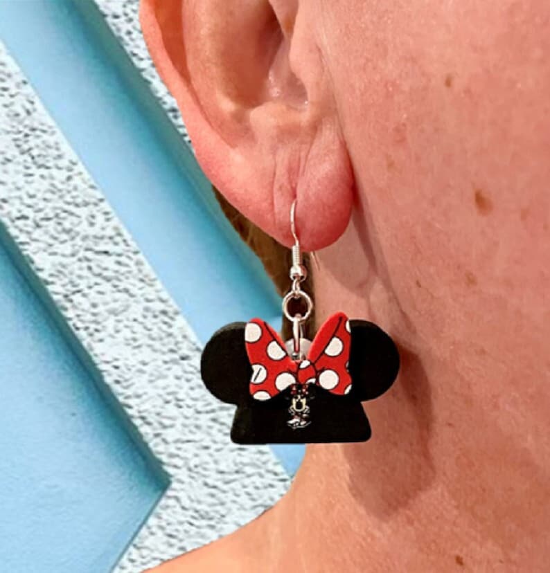 May include: A pair of black Minnie Mouse-themed earrings. Each earring features a black silhouette of Minnie's head with a red bow adorned with white polka dots. The earrings hang from silver-colored fish hook ear wires.