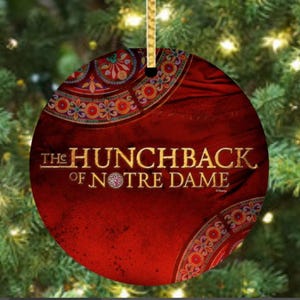 May include: Round Christmas ornament with the text "The Hunchback of Notre Dame" on a red background. The ornament has a gold ribbon for hanging and is decorated with a colorful floral design.