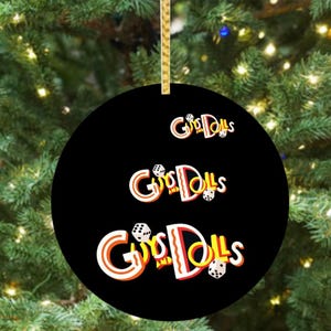 May include: A black round Christmas ornament with the words "Guys and Dolls" in a stylized font. The text is white with red and orange accents, and dice are included in the design. The ornament hangs from a gold ribbon.