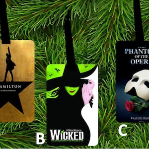May include: Three rectangular ornaments with black ribbons. One features a silhouette of a man and the text "HAMILTON AN AMERICAN MUSICAL" on a gold background. Another has a green witch and the text "WICKED A NEW MUSICAL". The third shows a white mask and the text "PHANTOM OF THE OPERA".