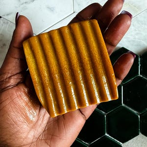 May include: A rectangular bar of soap with a vibrant mustard yellow color and a textured surface featuring deep, parallel vertical ridges. The soap has a slightly glossy finish and a dense, solid appearance, showcasing a uniform golden hue throughout.