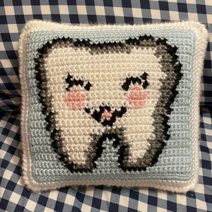 May include: A handmade crochet pillow in the shape of a smiling tooth. The tooth is white with gray and black accents, pink cheeks, and black eyes and mouth. The pillow has a light blue border and white trim.