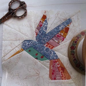 May include: A colorful fabric hummingbird quilt block with a floral background. The bird features blue, pink, orange, and green patterned wings and body. A pair of decorative scissors and a spool of thread are in the background.