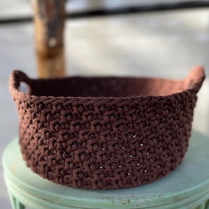 May include: A handcrafted, round crochet basket in a rich, dark brown color. The basket features two small handles and a textured stitch pattern. This decorative basket is suitable for storage or display.