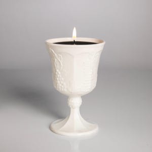 Puede incluir: Una vela blanca encendida en una copa decorativa. La copa tiene un tallo y un cuenco ancho y texturizado con un diseño de uvas. La cera de la vela es negra y la llama es amarilla y naranja.